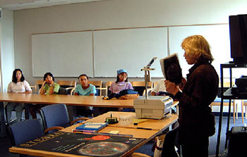 Janet Luhmann at the right holding up a book in a classroom. There are kids sitting in the background