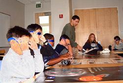 A few kids sitting at a table facing right. Some of them are putting on stere-vision glasses.