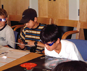 One student looking down with a pair of stereo-vision glasses at some poster. Another student is bending his stereo-vision goggles.