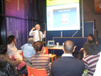 A presenter talking with a projected powerpoint presentation. A group is sitting around him, listening.