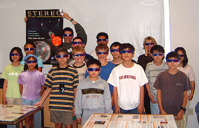 Sixteen kids stand in front of a classroom wearing stereo-vision glasses.